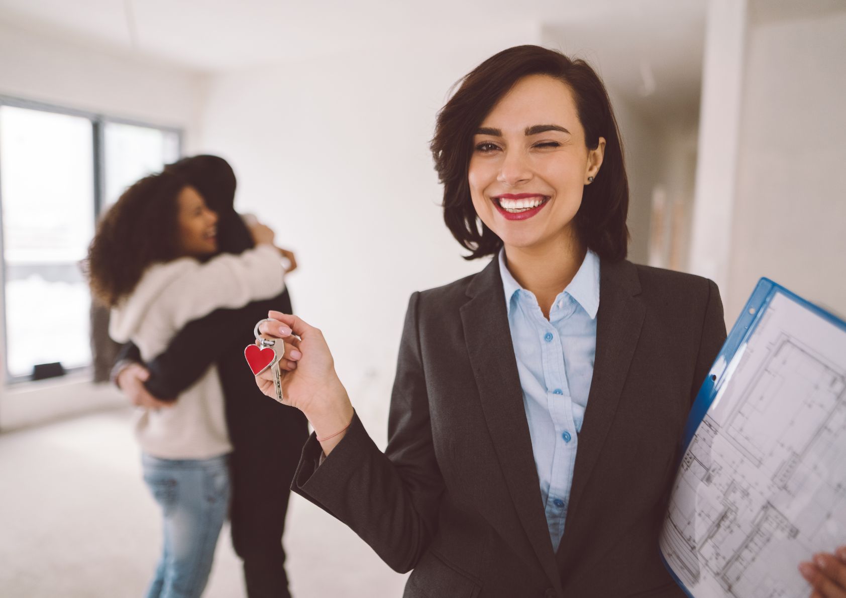 real estate agent holding keys while happy clients are hugging behind her. 